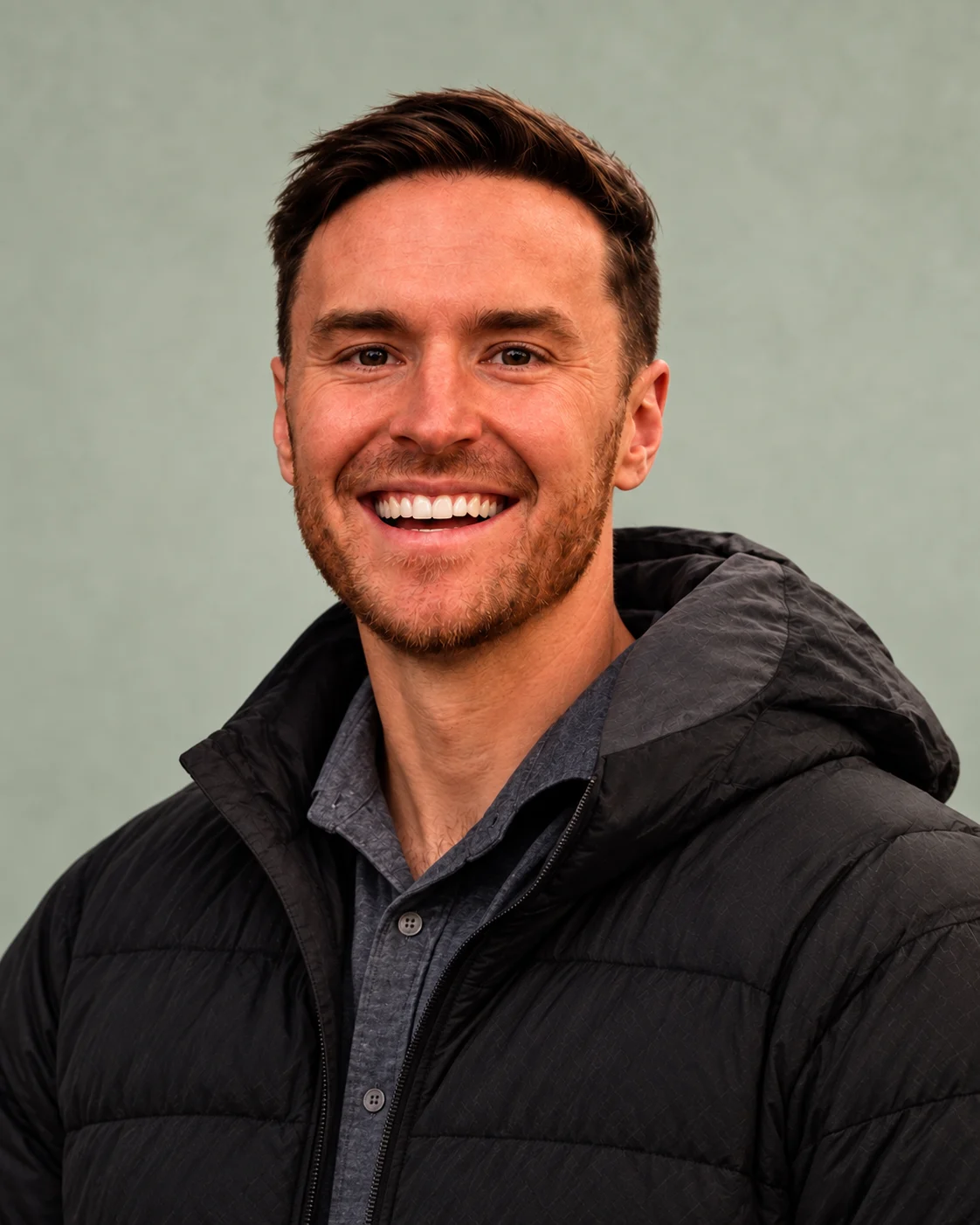 John Pollard smiling, wearing a charcoal jacket against a soft sage background.